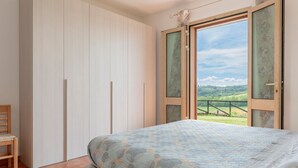A bedroom with a bed, a chair, a wardrobe, and a large window with a view of a green landscape. - Poggio Novello - Agriturismo Peccioli (Peccioli)