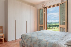 Un dormitorio con cama, una silla, un armario y un ventanal con vistas a un paisaje verde.