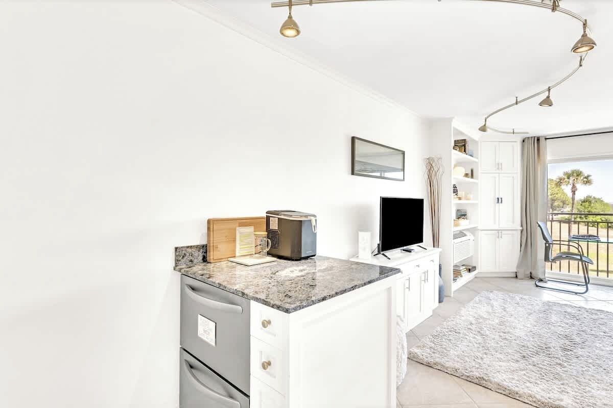Your bright kitchenette with granite counters and modern appliances opens to a cozy living space with balcony views of palm trees.