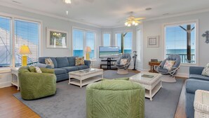 Een woonkamer met een grote televisie, groene en blauwe banken, rieten meubels en uitzicht op de oceaan.