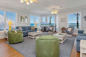 Ein Wohnzimmer mit großem Fernseher, grünen und blauen Sofas, Korbmöbeln und Blick aufs Meer.