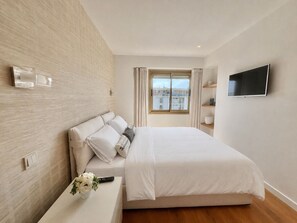 Ein modernes Schlafzimmer mit Bett, Fernseher, Fenster mit Vorhängen und Blick auf Gebäude.