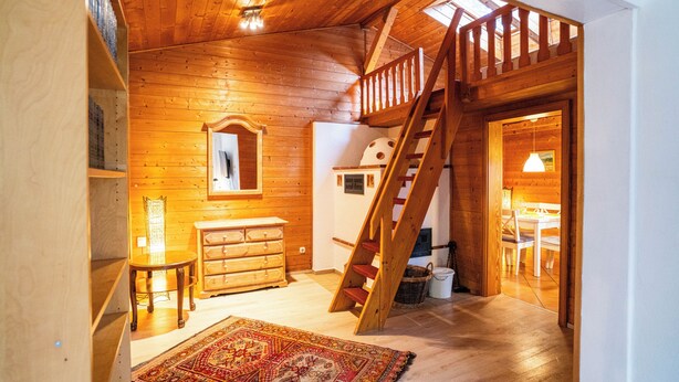 A wooden interior with a staircase, a dresser, a chair, and a patterned rug.