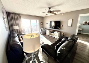 Ein Wohnzimmer mit Deckenventilator, Fernseher, einer Couch, einer Lampe und Blick ins Freie.