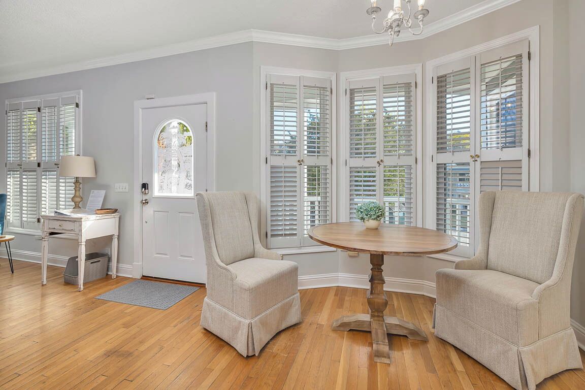Une pièce avec un plancher en bois, une porte blanche, un bureau blanc avec une lampe, deux fauteuils beiges, une table ronde et une fenêtre avec des volets.
