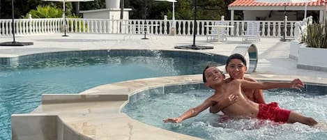 Una piscina con zona de hidromasaje, rodeada de palmeras y sombrillas.