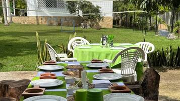 Un área de comedor al aire libre con una mesa preparada para cuatro personas, rodeada de sillas y una casa al fondo.