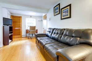A living room with a leather sofa, wooden flooring, a TV on a stand, a desk with a chair, and two framed pictures on the wall.