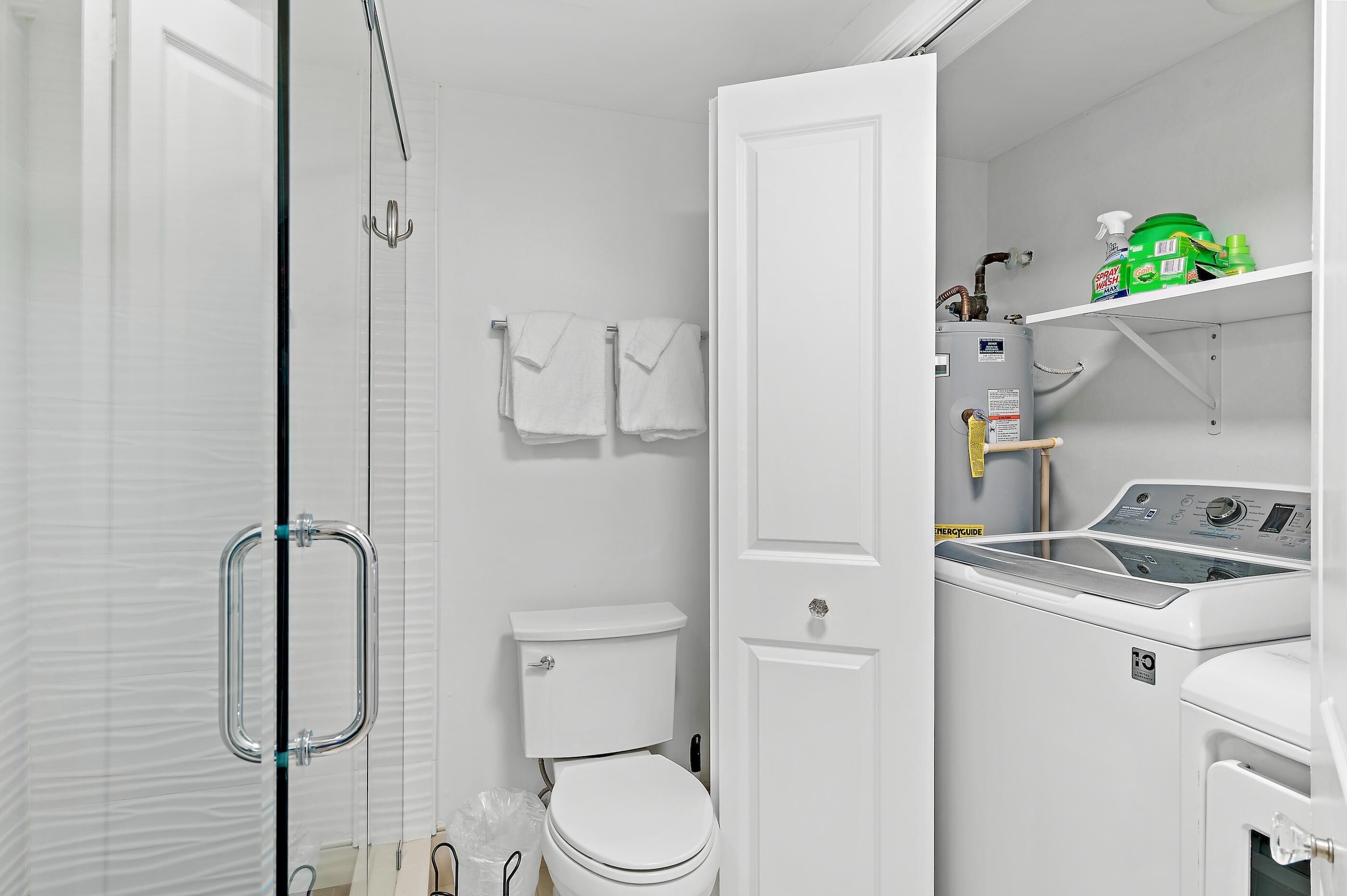 A bathroom with a glass shower enclosure, a toilet, a washing machine, and a shelf with cleaning supplies.