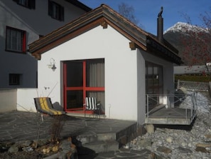 A white building with a red-framed door, a small porch with a chair, and a stone pathway leading to a staircase. - Baeder, (Müstair) by Interhome (Müstair)