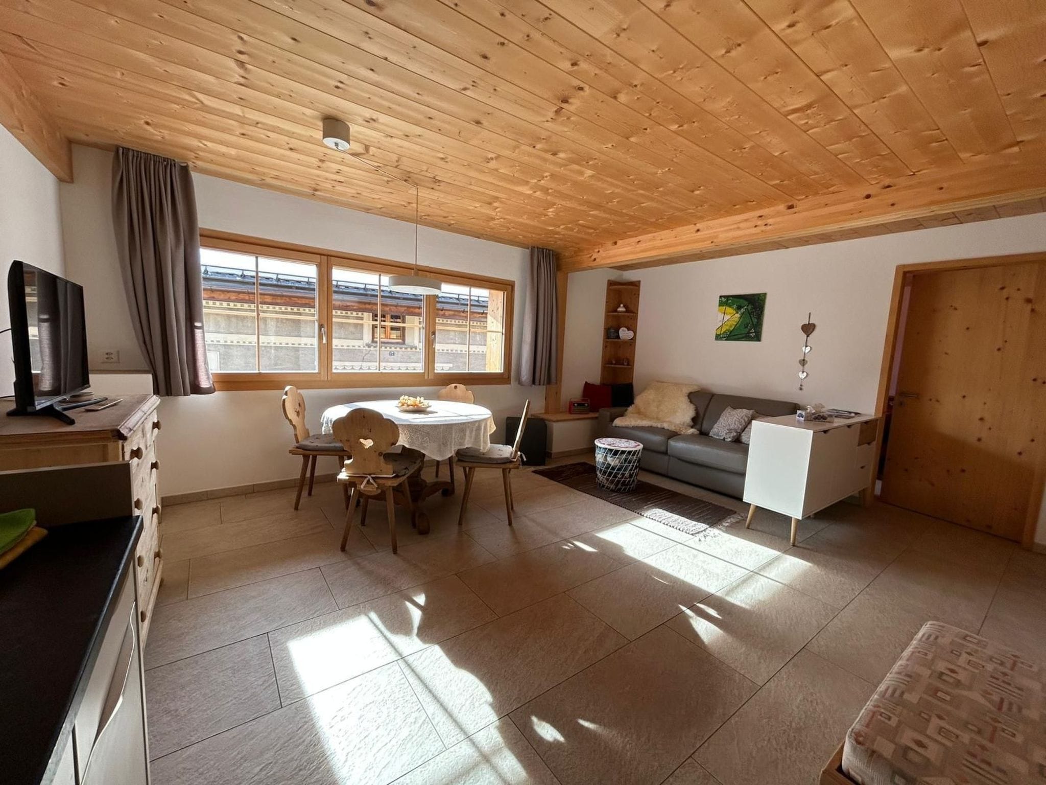 A spacious living area with a wooden ceiling, a dining table with chairs, a sofa, a TV, and a large window with curtains.