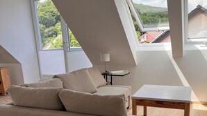 Camera da loft moderna con soffitto spiovente, un divano beige, un tavolino e vista su verde e edifici attraverso ampie finestre.