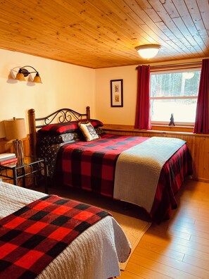 Quarto com duas camas, paredes de madeira, janela com cortinas, abajur e um quadro pendurado na parede.