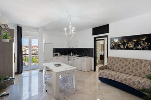 Een moderne keuken met een centraal eiland, een eethoek met een tafel en stoelen, een bank en een groot raam met uitzicht op gebouwen.