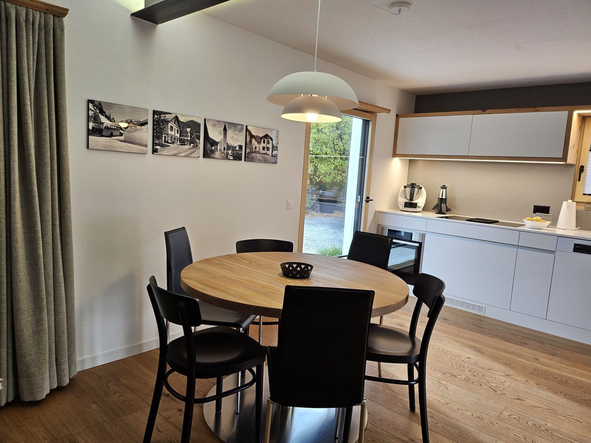 A modern kitchen with a dining area, featuring a round wooden table, black chairs, and a pendant light.