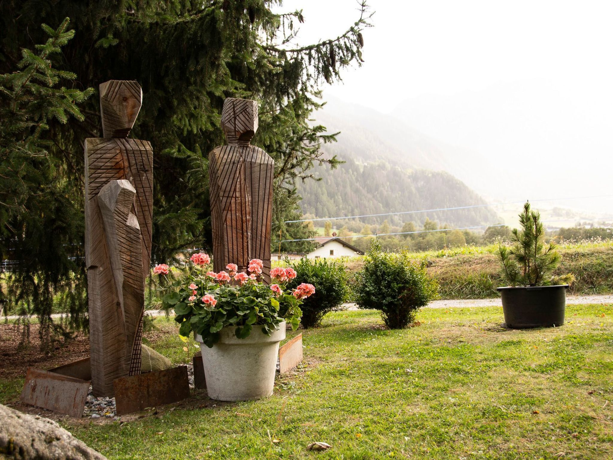 Two wooden sculptures in a garden with a large potted plant of pink flowers and a small tree.