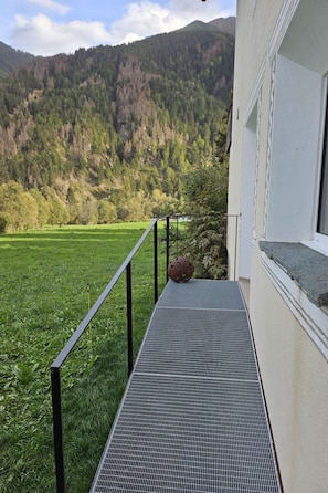 Un balcone con pavimento a griglia di metallo, ringhiera nera e vista su un campo verde e colline boscose.