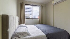 Een slaapkamer met een bed, een raam met gordijnen, een airconditioning en uitzicht op gebouwen.