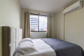 Ein Schlafzimmer mit Bett, einem fensterbelaubten Fenster, einer Klimaanlage und Blick auf Gebäude.