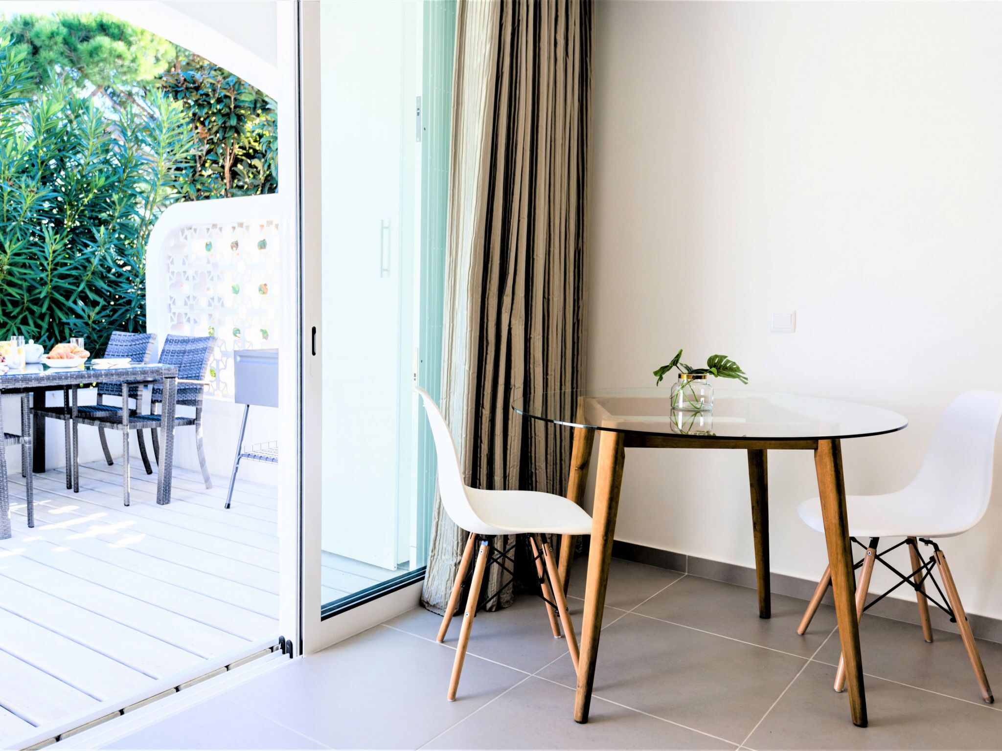 Un espace repas moderne avec une table et des chaises en verre, une vue sur la verdure à l’extérieur et un rideau à la fenêtre.