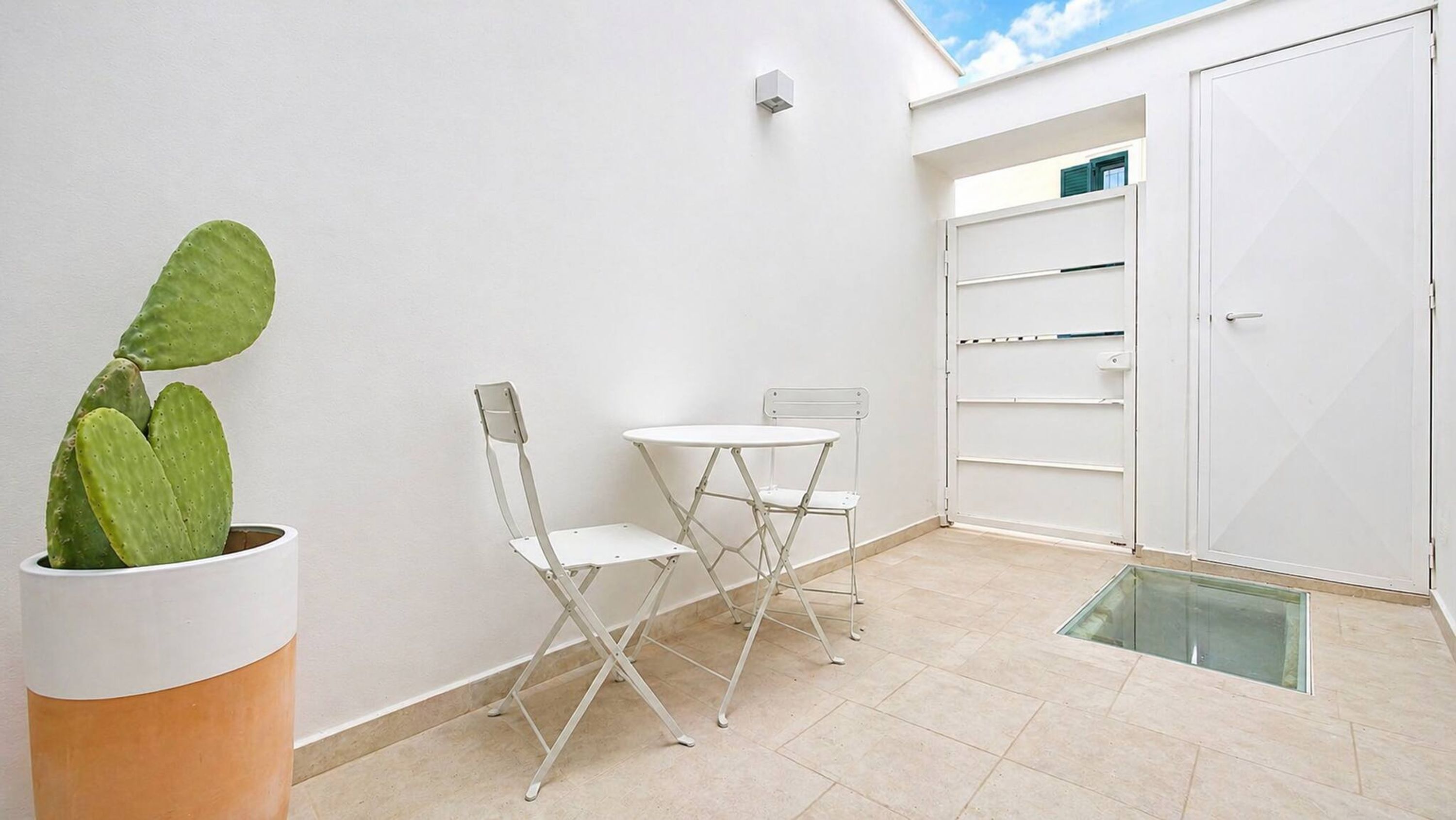 Una habitación pequeña y limpia con una puerta blanca, una mesa y silla blancas, una planta en maceta y una puerta de cristal que da al exterior.