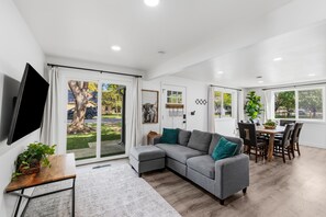 A modern living room with a sectional sofa, dining table, and chairs, large windows, and a flat-screen TV.