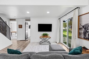 A modern living room with a flat-screen TV, grey sofas, a wooden coffee table, and a large window with a view of a green landscape.
