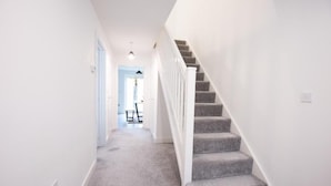 Ein Flur mit Teppichboden, einer weißen Treppe und einer Tür zu einem Raum mit Tisch und Stuhl.