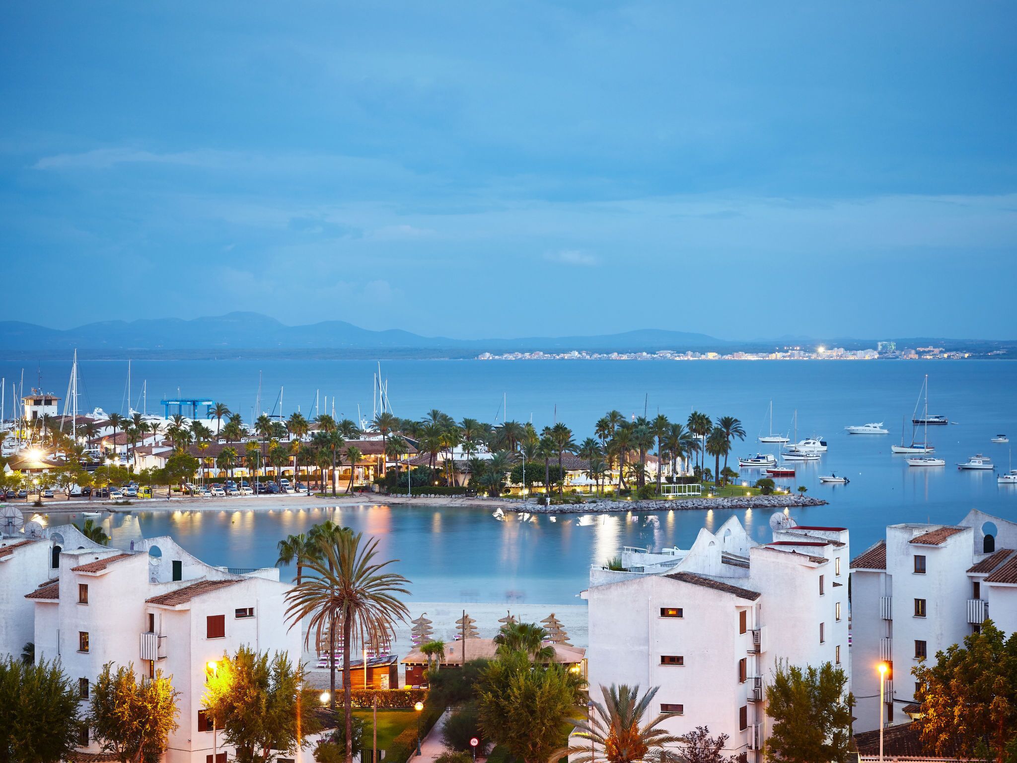 A coastal town with white buildings, a marina with boats, and a view of the sea and distant cityscape.