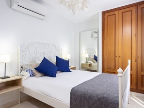 A bedroom with a white bed, two blue pillows, a bedside table with a lamp, a wardrobe, and an air conditioning unit. - TEBAR BEACH by Interhome (Playa Blanca)