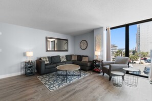 Un salon moderne avec un canapé d’angle, une table basse ronde, une petite table d’appoint et un fauteuil. Une grande fenêtre offre une vue sur la ville.