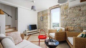 Un salon avec un canapé, une table d’appoint rouge, un fauteuil en osier, un téléviseur posé sur un meuble blanc et un mur en pierre.