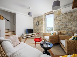 A living room with a sofa, a red side table, a wicker chair, a television on a white stand, and a stone wall.