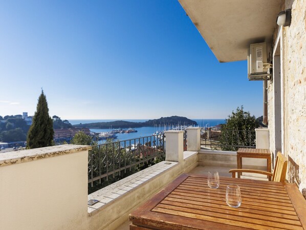 A balcony with a wooden table and two glasses, overlooking a harbor with boats and a distant island.