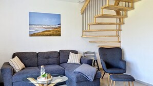 Ein Wohnzimmer mit grauem Sofa, einem Couchtisch, einem Sessel, einer Holzspirale und einem Gemälde einer Strandansicht.