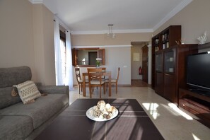 Ein Wohnzimmer mit einer Couch, einem Esstisch mit Stühlen, einer TV-Konsole und einer Küchenzeile im Hintergrund.