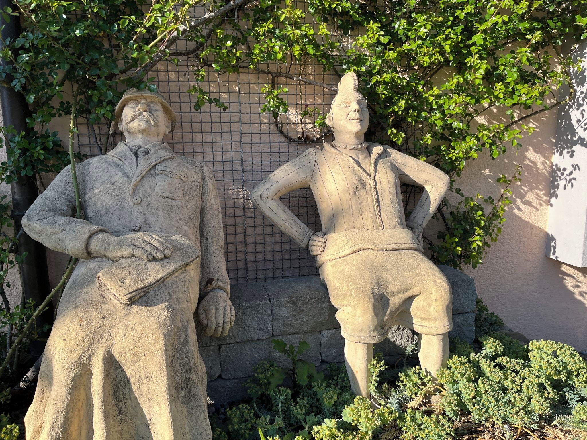 Two stone statues in an outdoor setting with a brick wall and greenery in the background.