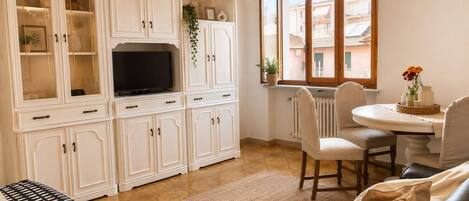 Una sala de estar con televisor, armarios blancos, una mesa de comedor con sillas y una ventana con vista.