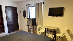 Una habitación con una cama, un televisor montado en la pared, una mesita con una silla y una ventana con cortinas.