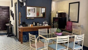 Un comedor con una mesa redonda, sillas blancas, piso con estampado azul y blanco, un armario de madera, un espejo y un ventilador.