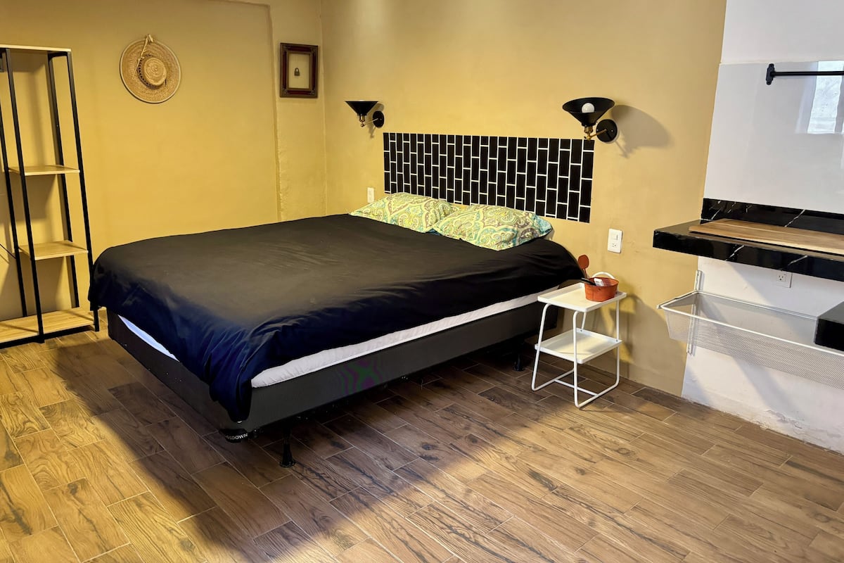 A bedroom with a bed, a nightstand, a wall-mounted shelf, a ceiling fan, and a window.