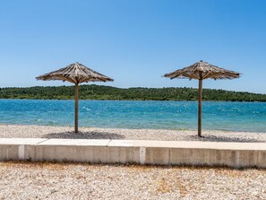 Zwei strohgedeckte Sonnenschirme an einem Kiesstrand mit blauem Meer und einer bewaldeten Hügelkette im Hintergrund.