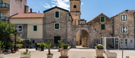 A historic stone building with a clock tower, arched entrance, and a paved square with potted plants.