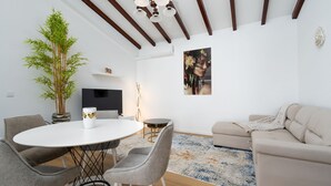 En moderne stue med et rundt spisebord, grå stole, en beige sofa, en stor stueplante, et fladskærms-TV og et billede i ramme på væggen.