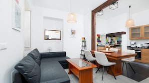 A modern living room with a black leather sofa, a wooden coffee table, and a dining area with a wooden table and chairs. There is a kitchen visible in the background with colorful tiles and wooden cabinets. - Charming Garden Oasis by LovelyStay (Funchal)
