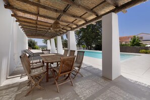 An outdoor patio with a wooden table and chairs, a pool in the background, and a clear sky.