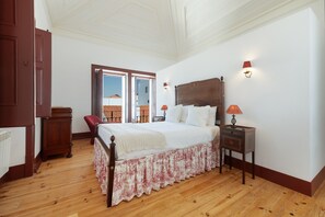 A bedroom with a wooden floor, a bed with a white bedspread, a red chair, a nightstand with a lamp, and a view through an open door.