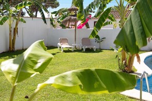 A poolside area with a grassy lawn, lounge chairs, and tropical plants.
