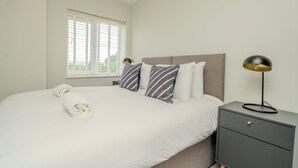 A neatly made bed with a rolled towel, a bedside table with a lamp, and a window with blinds. - Church House by Guestz (Filton)