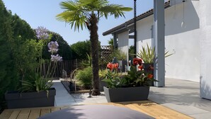 A patio with a wooden deck, a grey cushion, potted plants, and a palm tree. - 5-Room Villa with Pool and Terrace in Le Porge (Le Porge)